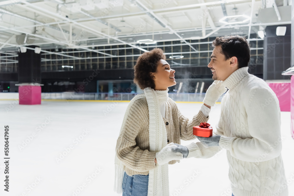 Side view of smiling man holding heart shaped gift box and hand of african american girlfriend on ice rink