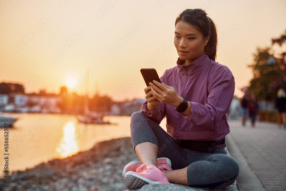 Asian sportswoman using smart phone while relaxing outdoors at sunset.