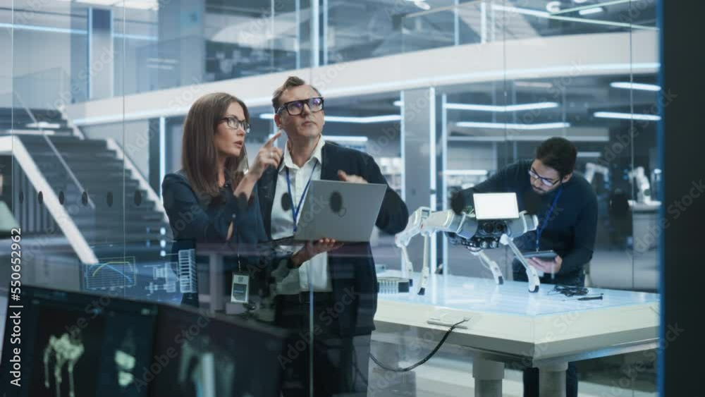 Chief Technology Officer Talking to Project Manager, Discussing Robot Dog Concept in High Tech Research and Development Facility. Professional Industrial Science Specialists Working in the Background. Chief Technology Officer Talking to Project Manager, Discussing Robot Dog Concept in High Tech Research and Development Facility. Professional Industrial Science Specialists Working in the Background.
