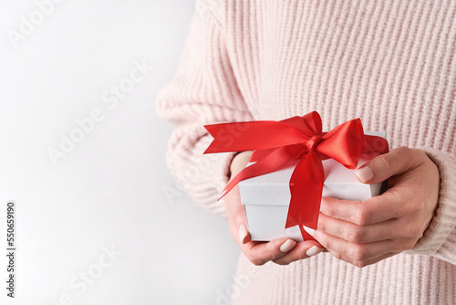 Fototapeta Naklejka Na Ścianę i Meble -  Female hands in woolen pink sweater holding white gift box with red ribbon on light background. Christmas, birthday or wedding composition with copy space. Mockup for New Year or Christmas.