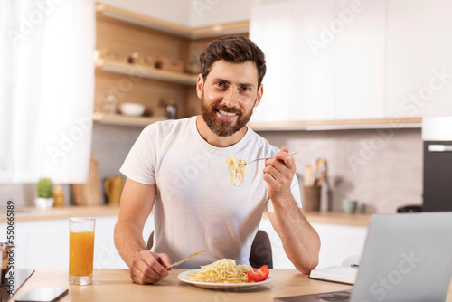 Happy handsome mature caucasian bearded male eating pasta in minimalist kitchen interior with laptop