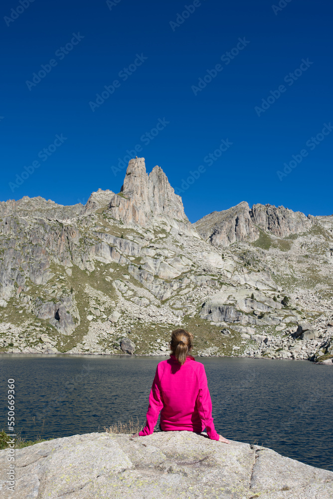Fototapeta premium A backpacker woman in a pink jacket sitting outdoors on a rock in front of the lake, contemplate the needles of the mountain. Epic vacations and adventure in the Aigüestortes National Park. Back view.