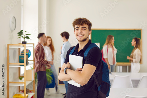 Portrait of happy caucasian student with blue backpack and big tablet computer against background of class with his classmates. College lifestyle, future professionals. Selective focus.