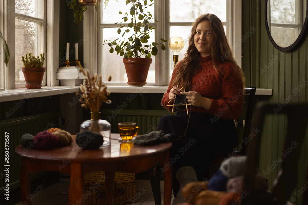 A young caucasian woman with long hair wearing a red knitted sweatshirt sitting in a chair in an old house knitting.