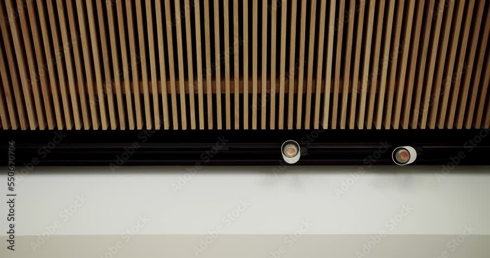 Decorative timber panel with lamp. Low angle pan left view of decorative lumber panel with lamps installed on ceiling in light room. Wooden panels in minimalist interior with modern design. Decorative timber panel with lamp. Low angle pan left view of decorative lumber panel with lamps installed on ceiling in light room. Wooden panels in minimalist interior with modern design.