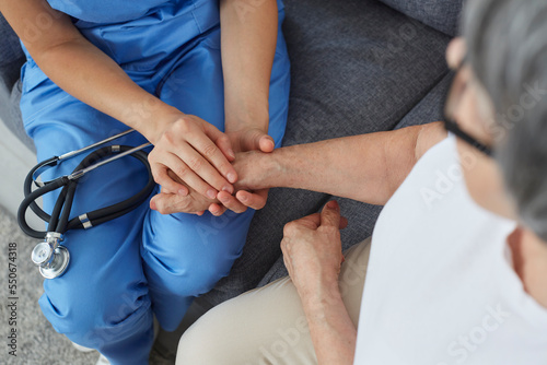 Doctor giving compassion to patient. Young physician or cardiologist in blue scrubs, with stethoscope on lap sitting on couch and holding senior woman patient by hand. Support concept. Cropped shot