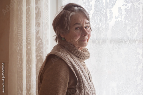 An elderly beautiful retired woman looks in the window with lace curtains.