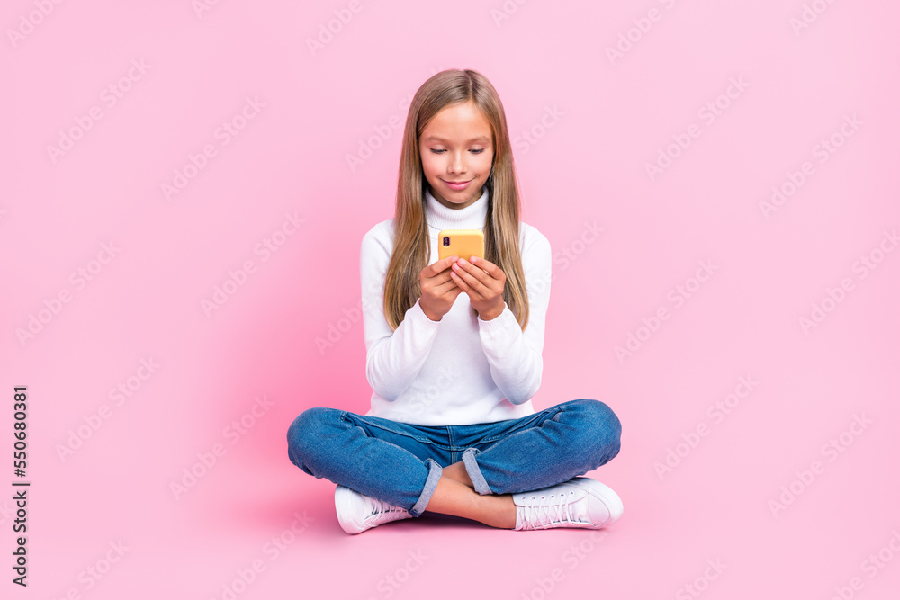 Full size portrait of cute positive schoolkid sitting floor use telephone isolated on pink color background Full size portrait of cute positive schoolkid sitting floor use telephone isolated on pink color background