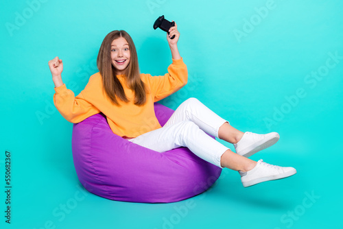 Full length photo of cute lucky school girl dressed orange hoodie bean bag winning playstation empty space isolated teal color background