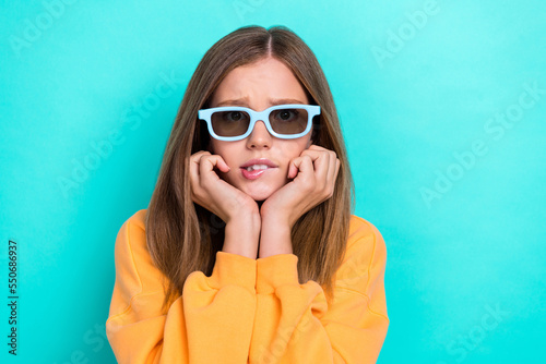 Photo of cute teenager girl brown hair touch cheeks nervous bite lips watching film wear glasses dramatic moment isolated on cyan color background