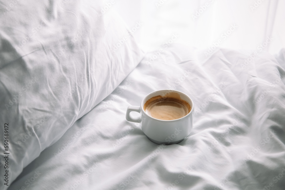 Cup of coffee in bed with white cotton bedclothes. Morning coffee concept. Sunday loneliness relaxing, hygge mood. Minimalism composition. Light and shadow. White mug of americano or expresso Cup of coffee in bed with white cotton bedclothes. Morning coffee concept. Sunday loneliness relaxing, hygge mood. Minimalism composition. Light and shadow. White mug of americano or expresso