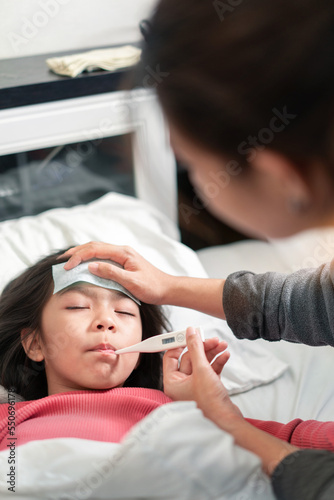 Asian mother measuring temperature girl and hand on forehead on bed at morning time, Sick child have cool gel for reduce high fever, Selective focus, Healthy and infection concept