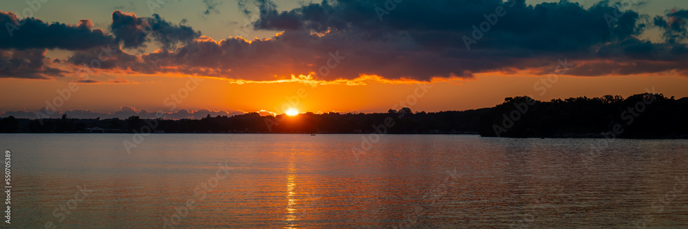 Serene sunset scene at a Minnesota lake with calm water and clouds and the sun setting behind trees on the distant shoreline.