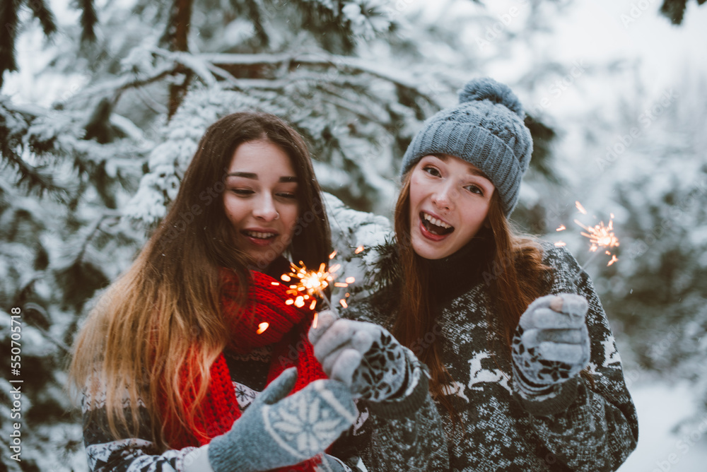 Close up fashion portrait of two sisters hugs and having fun, holding sparklers in winter time forest, wearing sweaters and scarfs,best friends couple outdoors, snowy weather Close up fashion portrait of two sisters hugs and having fun, holding sparklers in winter time forest, wearing sweaters and scarfs,best friends couple outdoors, snowy weather