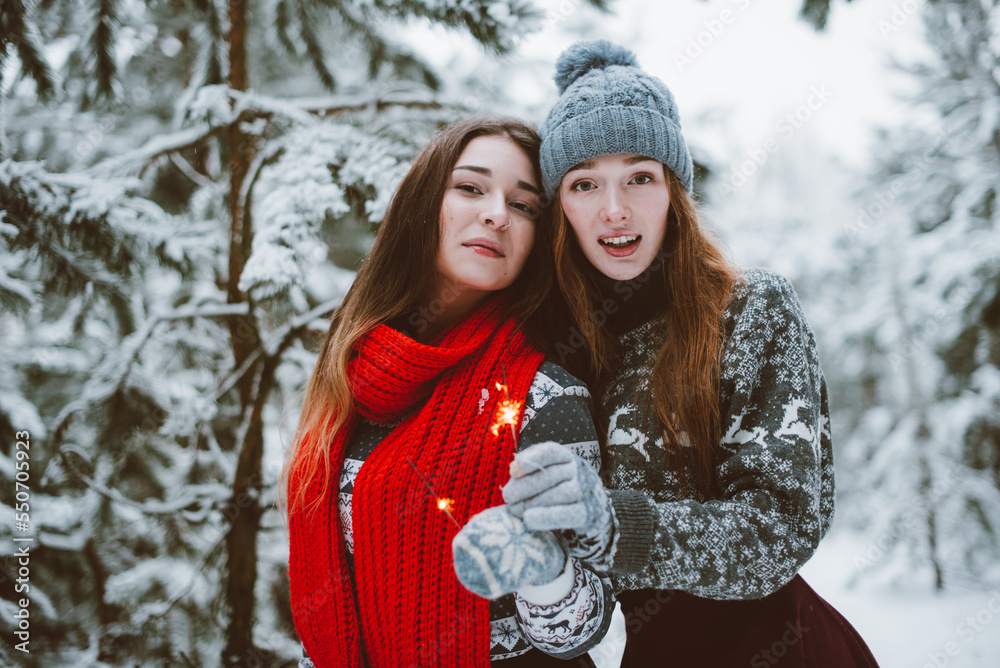 Fototapeta premium Close up fashion portrait of two sisters hugs and having fun, holding sparklers in winter time forest, wearing sweaters and scarfs,best friends couple outdoors, snowy weather