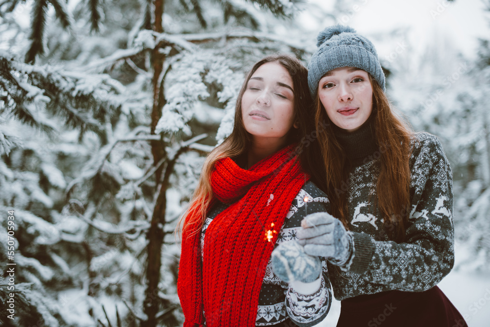 Fototapeta premium Close up fashion portrait of two sisters hugs and having fun, holding sparklers in winter time forest, wearing sweaters and scarfs,best friends couple outdoors, snowy weather