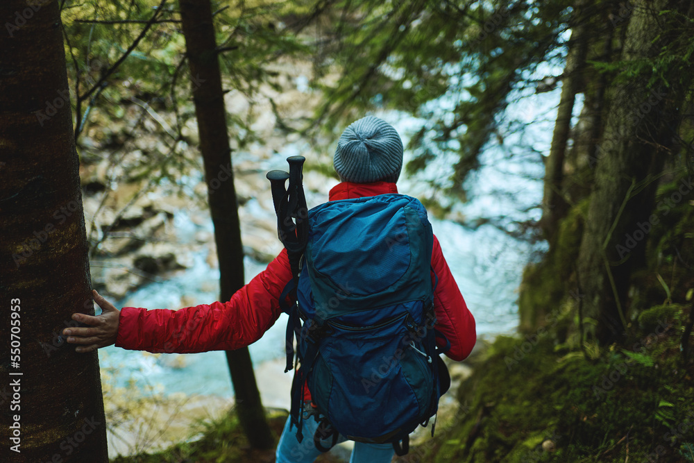 Back view of woman hiker with backpack resting by the forest blue river. Travel adventure hiking with backpack outdoors