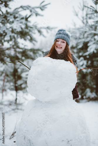 young teenage hipster girl having fun, make snowman in winter time forest, wearing sweaters and scarfs, outdoors, snowy weather