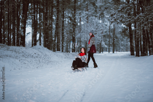 Close up fashion portrait of two sisters hugs and having fun, ride on sled in winter time forest, wearing sweaters and scarfs,best friends couple outdoors, snowy weather