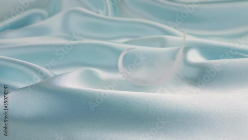 White swan feathers fall on pale blue pastel silk. Slow motion.