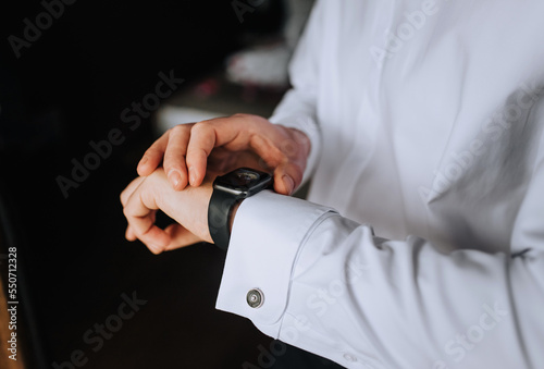A man  a businessman in a white shirt holds a smartwatch  a wristwatch on his hand  preparing in the morning. Close-up photography  business.