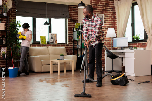 Happy african american man vacuuming floor while concentrated woman dusts decorative objects. Interracial couple cleaning home in harmony, doing household chores together.