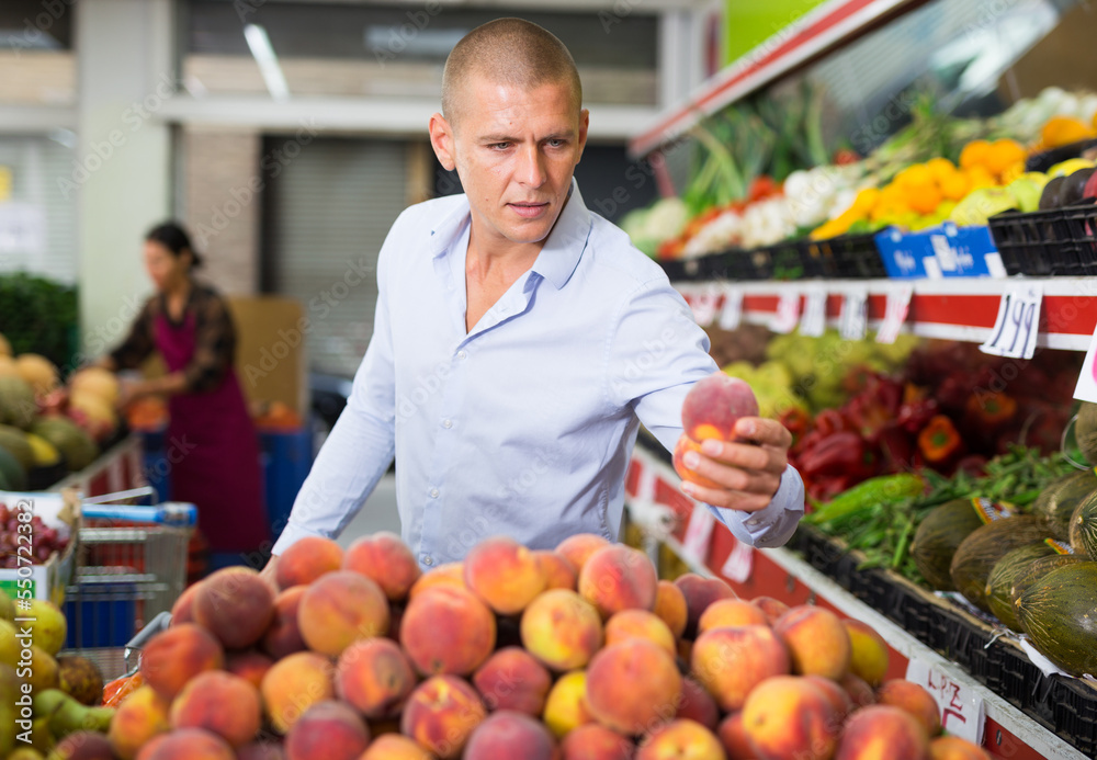 Portrait of positive adult man choosing fresh ripe peaches from counter at farmers market