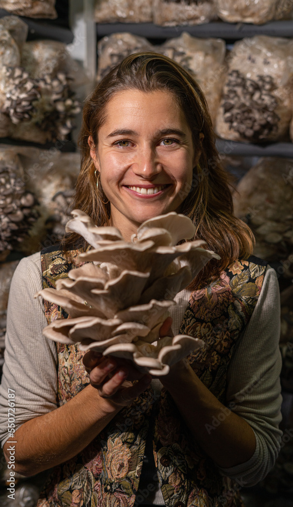 happy young woman smiling to camera and holding mushrooms against the oyster mushroom farm. growing up cultivated mushrooms at home