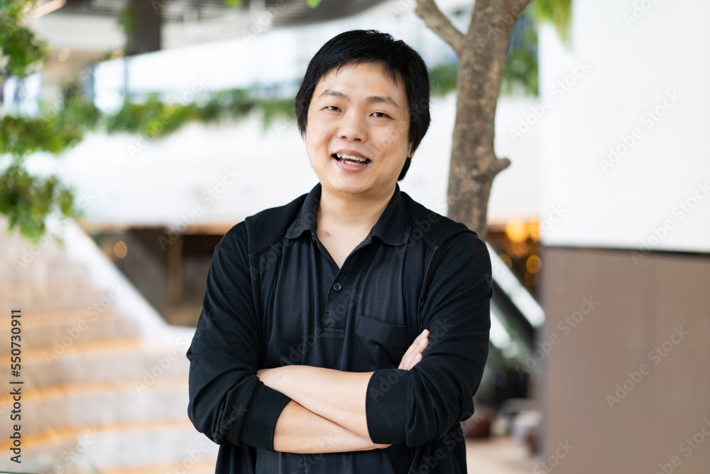 The portrait of Asian man in a black casual shirt with crossed arms standing in front of a blurred background of an office workspace. The portrait of Asian man in a black casual shirt with crossed arms standing in front of a blurred background of an office workspace.