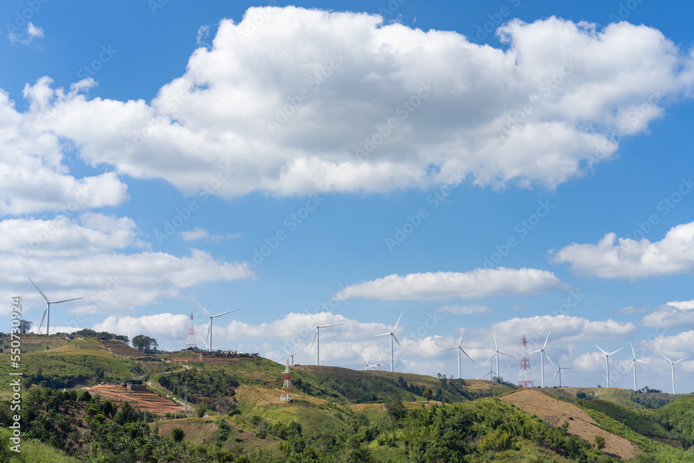 The wind turbines in the top of the grass hill underneath the group of white clouds and blue sky on a sunny day, produced electricity from pure energy.