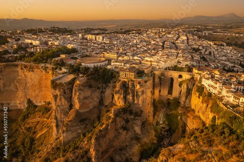 Aerial photo of Ronda, Spain, at sunset. The hilltop village is bathed in golden light. Bullring and Puente Nuevo bridge are visible.