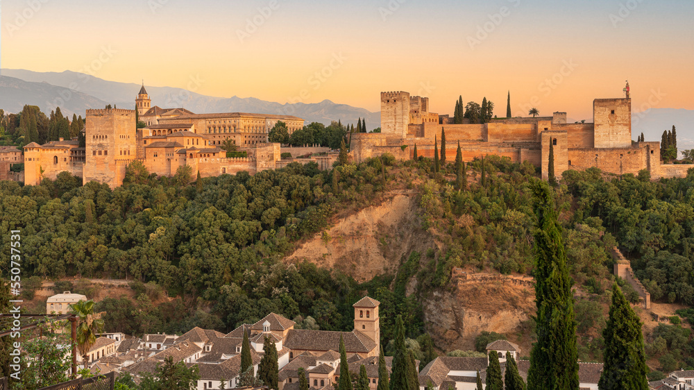 Sunset photo of the Alhambra fortress in Granada, Spain. The fortress is bathed in a golden, reddish glow.
