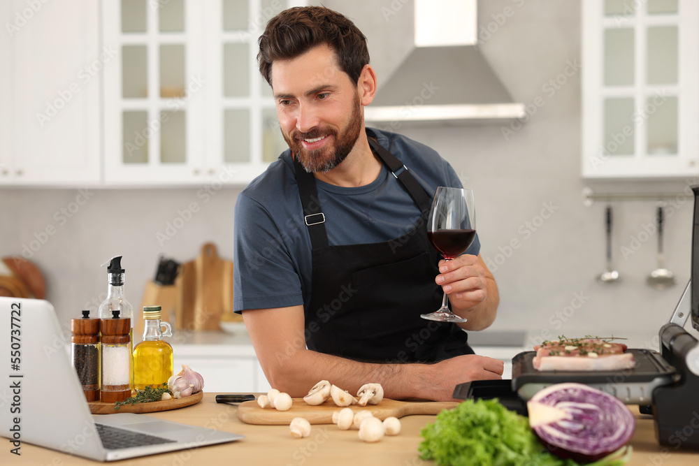 Man with glass of wine making dinner while watching online cooking course via laptop in kitchen Man with glass of wine making dinner while watching online cooking course via laptop in kitchen
