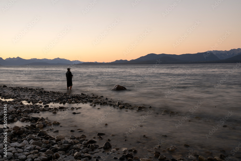 Night shot. View the beautiful water blurred effect and blurred fisherman dark silhouette, fishing at nightfall. The rocky shore and mountains in the horizon with a beautiful dusk light.