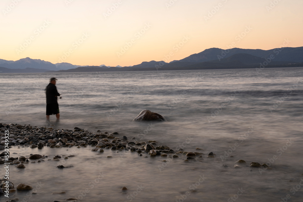 Night shot. View the beautiful water blurred effect and blurred fisherman dark silhouette, fishing at nightfall. The rocky shore and mountains in the horizon with a beautiful dusk light.