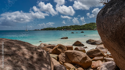 Seascape on a tropical island. A pile of boulders on a sandy beach. The yacht  in the turquoise ocean. A green hill against a blue sky  clouds. Granite large stones in the foreground. Seychelles