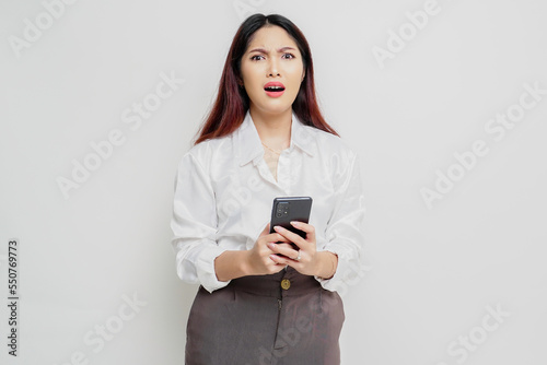 A dissatisfied young Asian woman looks disgruntled wearing white shirt irritated face expressions holding her phone