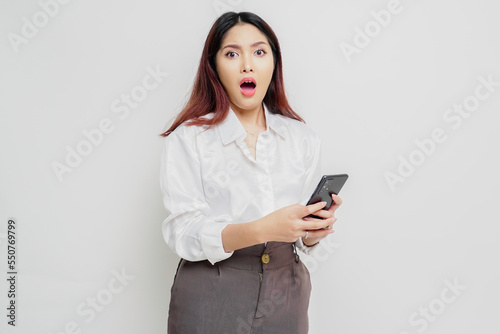 A dissatisfied young Asian woman looks disgruntled wearing white shirt irritated face expressions holding her phone