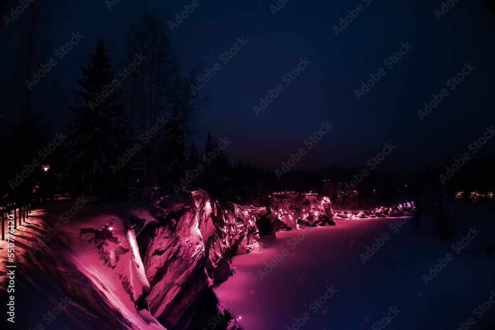 Beautiful panoramic view of night park with colorful lights Viva Magenta on winter forest and rocks, Ruskeala, Karelia, Russia. Trendy creative design in color of 2023. High quality photo