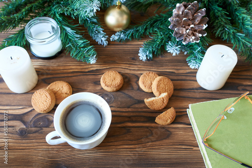 A coffee mug, cookies, and a book with glasses against the backdrop of Christmas tree branches. The concept is New Year's vacations, time to read books.