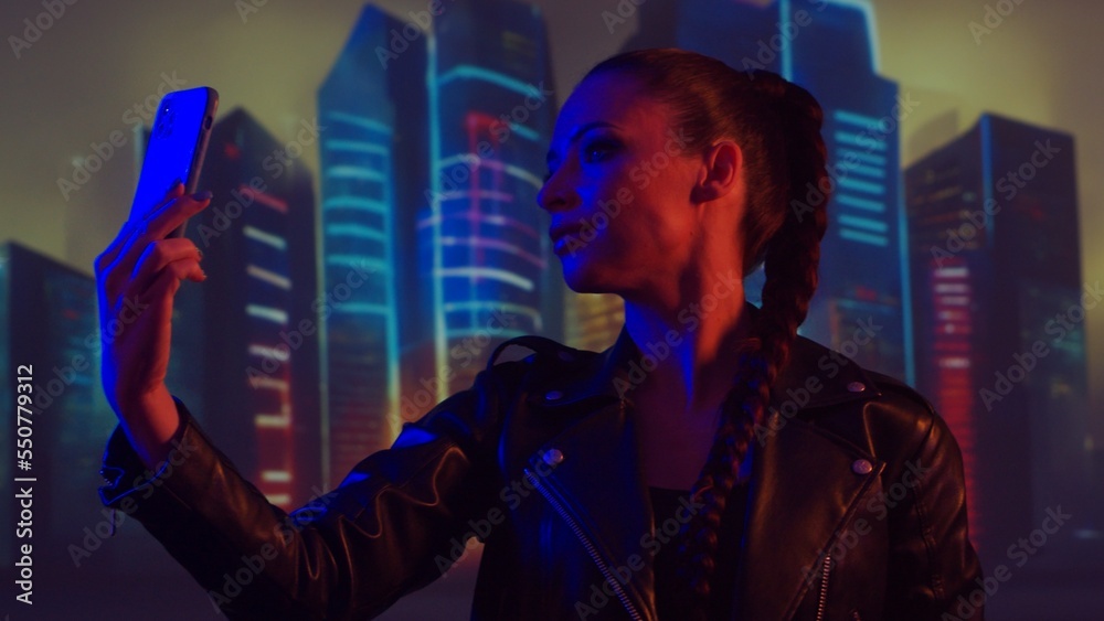 Fototapeta premium Portrait of cyberpunk girl with a smartphone. Beautiful young woman on the background of city scyscrapers. Futuristic concept.