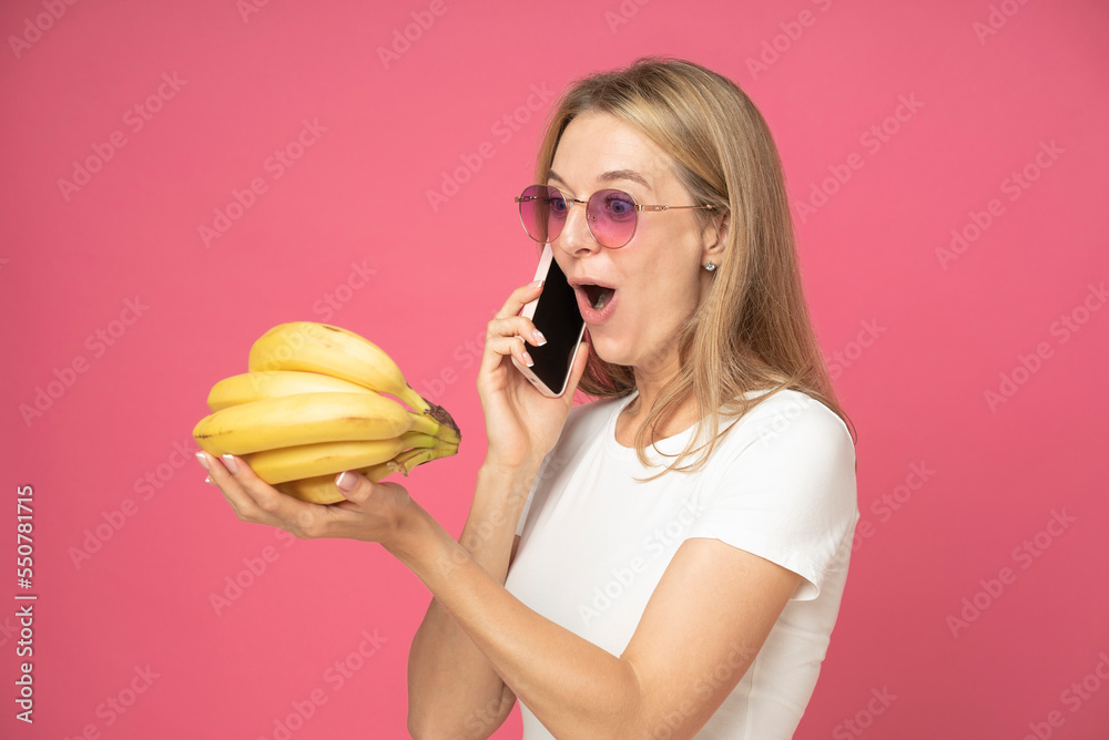 Shocked surprised pretty woman dressed in white t-shirt talking on mobile phone and looking on bananas on her hand isolated over pink colored background Shocked surprised pretty woman dressed in white t-shirt talking on mobile phone and looking on bananas on her hand isolated over pink colored background