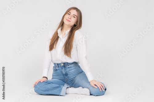Beautiful long-haired girl in a white shirt and blue jeans sits on the floor on a white background