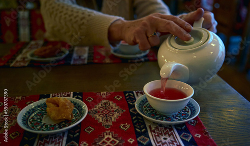Anonymous woman pours tea from a white ceramic teapot into a cup and paklava in a plate on a restaurant table. Restaurant with Crimean Tatar cuisine.