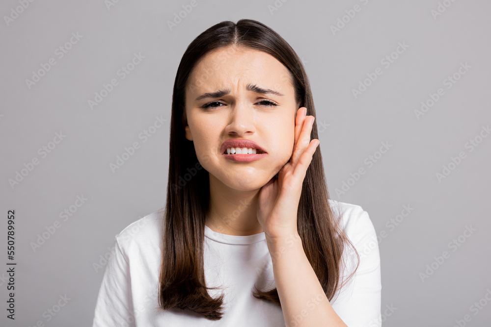 Portrait of crying sad tired woman lady standing on grey background wearing basic white t-shirt holding hand near head showing painful expressions suffering from tooth pain dental illness. Portrait of crying sad tired woman lady standing on grey background wearing basic white t-shirt holding hand near head showing painful expressions suffering from tooth pain dental illness.