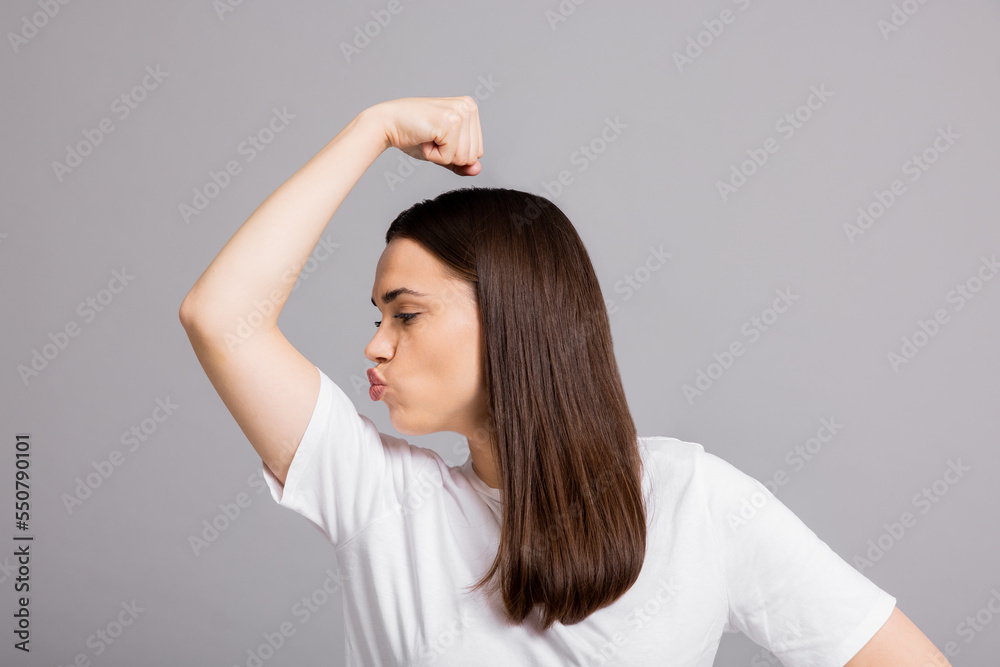Successful young self made lady girl woman raising hand showing biceps kissing her biceps success gesture over white background wearing basic white t-shirt. Successful young self made lady girl woman raising hand showing biceps kissing her biceps success gesture over white background wearing basic white t-shirt.