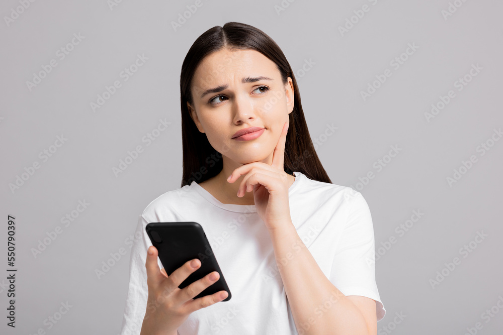Portrait of young cute successful cheerful woman thinkingshowing thoughtful expressions emotions girl in bad mood isolated on grey background holding cellphone showing delighted emotions at camera. Portrait of young cute successful cheerful woman thinkingshowing thoughtful expressions emotions girl in bad mood isolated on grey background holding cellphone showing delighted emotions at camera.