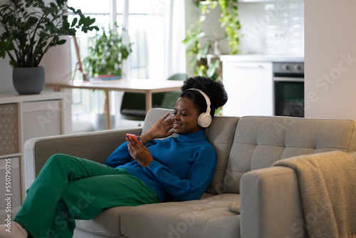 Happy African American woman in headphones listening to music using mobile phone to select playlist in popular online app. Smiling young girl lying on sofa of home and audition podcast or audiobook photo