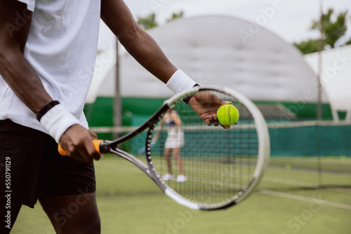 Close-up of the muscular hands of man of African appearance holding racket and tennis ball and preparing to serve. Boy is dressed in white T-shirt, black shorts and has wristbands on hands. © ABCreative