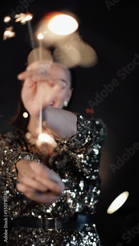 Vertical video. Young woman dancing with sparklers at christmas party on black background. Shiny brunette caucasian female with bengal lights. Glamour woman in golden dress.
